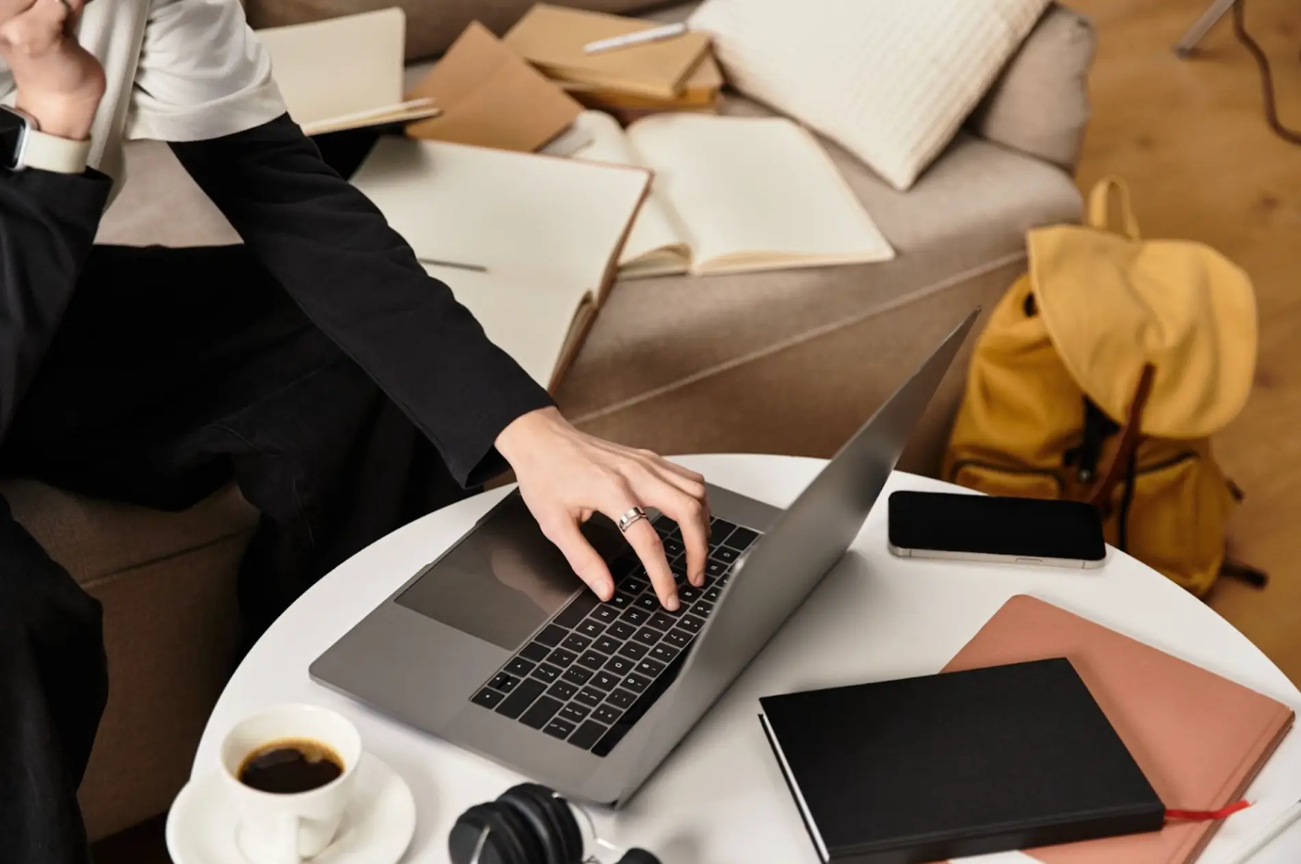 A person typing on a laptop surrounded by notebooks, coffee, and a smartphone, creating a cozy workspace ideal for planning email marketing for real estate agents. The scene suggests productive work in a home setting.
